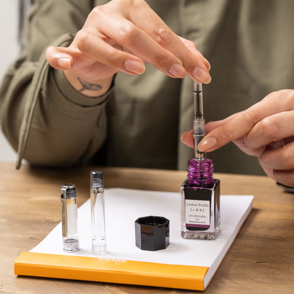 Pilot iroshizuku ink in fuschia purple, called yama-budo, which translates to Crimson Glory Vine in Japanese, being used to refill a Pilot Prera Transparent fountain pen, showcasing the environmental benefit of fountain pens.