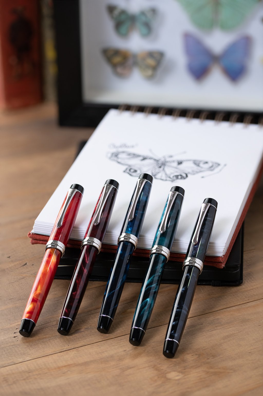 Pilot Custom Heritage SE fountain pens in five colours, leaning against a note pad with a butterfly drawing.